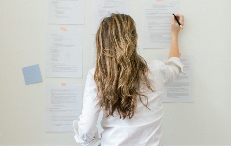 A woman sits on the floor in front of a wall, facing away from camera. She writes on pieces of paper taped to the wall.