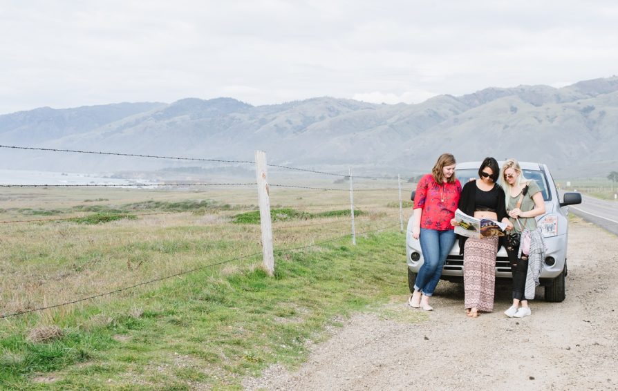 Three women stand in front of a car on the side of a road, reading from a magazine. In the distance are mountains.