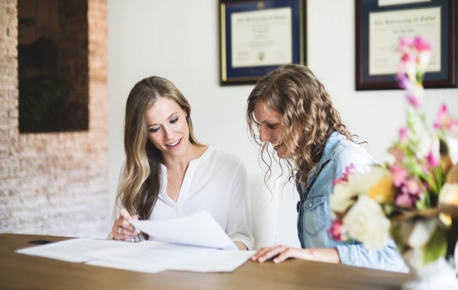 Two women sitting at a counter, glancing down at a group of papers and smiling.