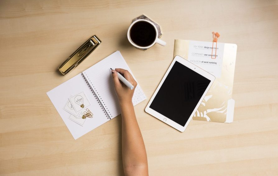 a desk with a person writing next to a tablet and cup of coffee