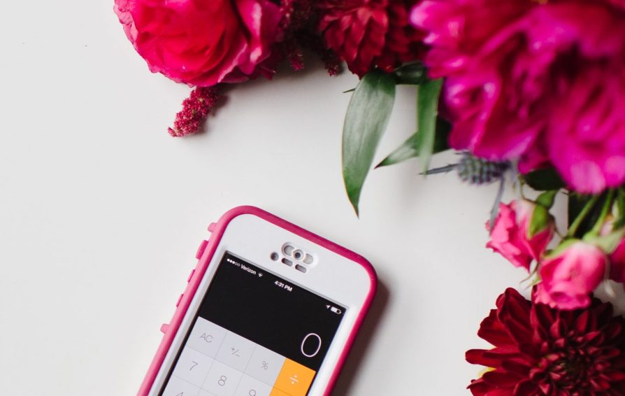 A phone with a pink case sits on a white surface with a calculator app open on the screen. The phone is framed by pink flowers.