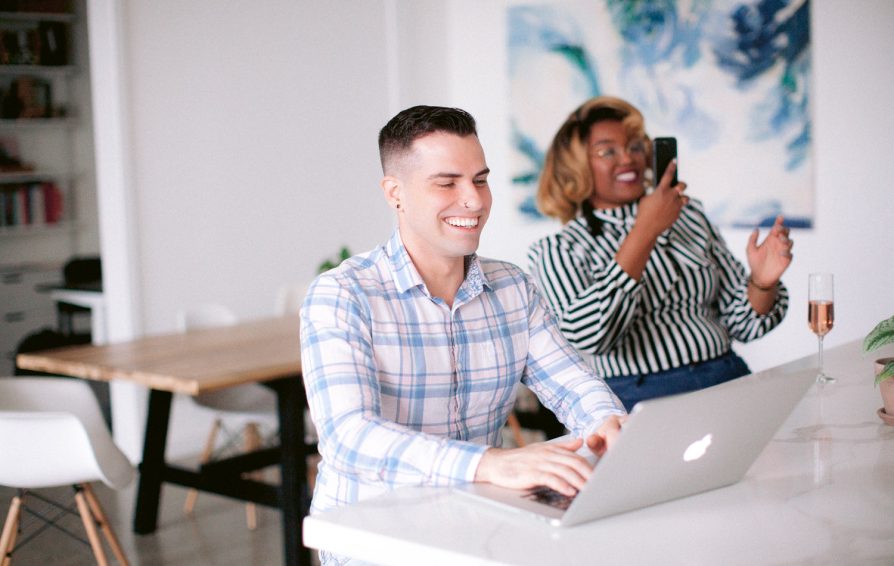 A man smiles, typing at a laptop. Beside him, a woman takes a picture on her phone