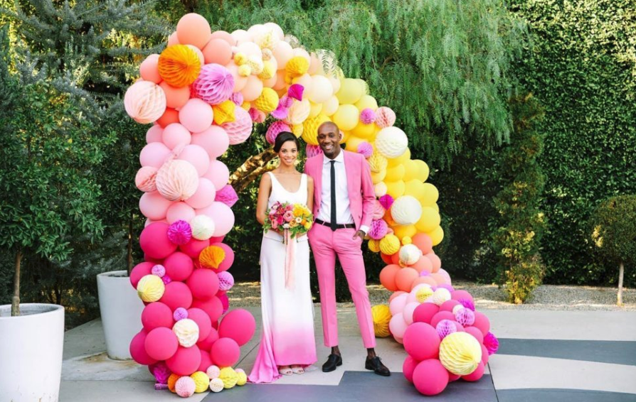 Bride and Groom in bright pink formal wear smile together under balloon arch.