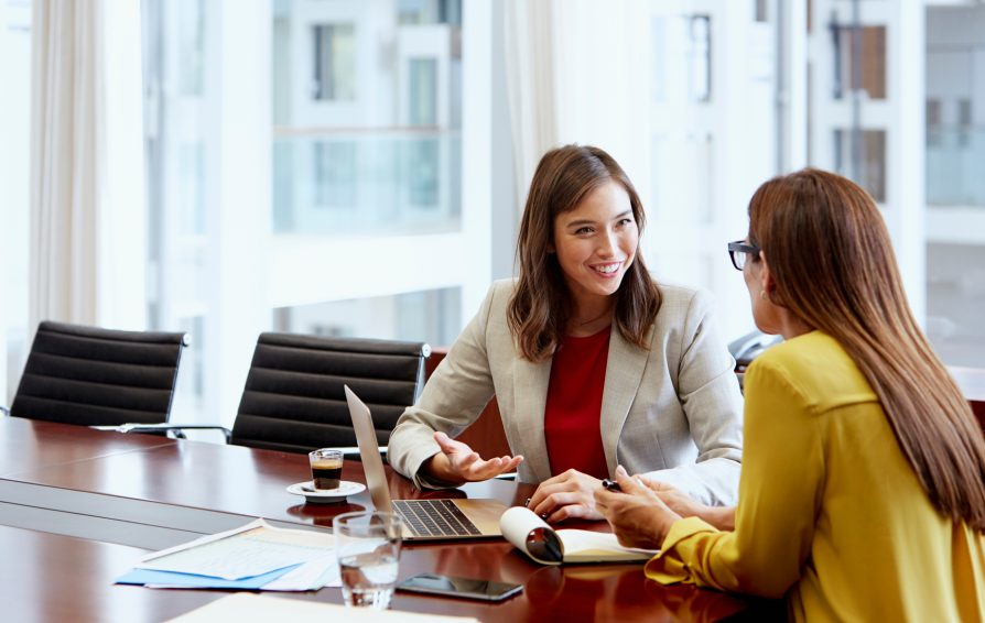Smiling businesswoman discussing with female colleague while sitting at conference table in brightly lit office