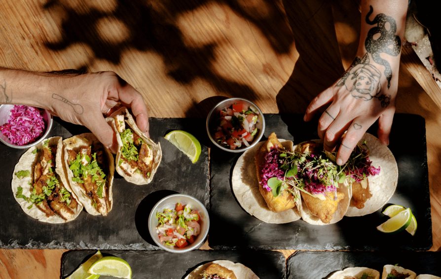 Assorted Mexican tacos on slates, on a wooden table in the sun, two hands with tattoos holding food. Picnic time