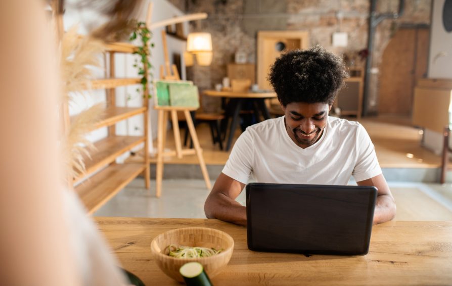 A man in a white t-shirt sits at a table, smiling down at his laptop.