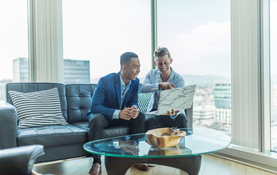 Two men in suits smile at a laptop in front of windows with a city view.