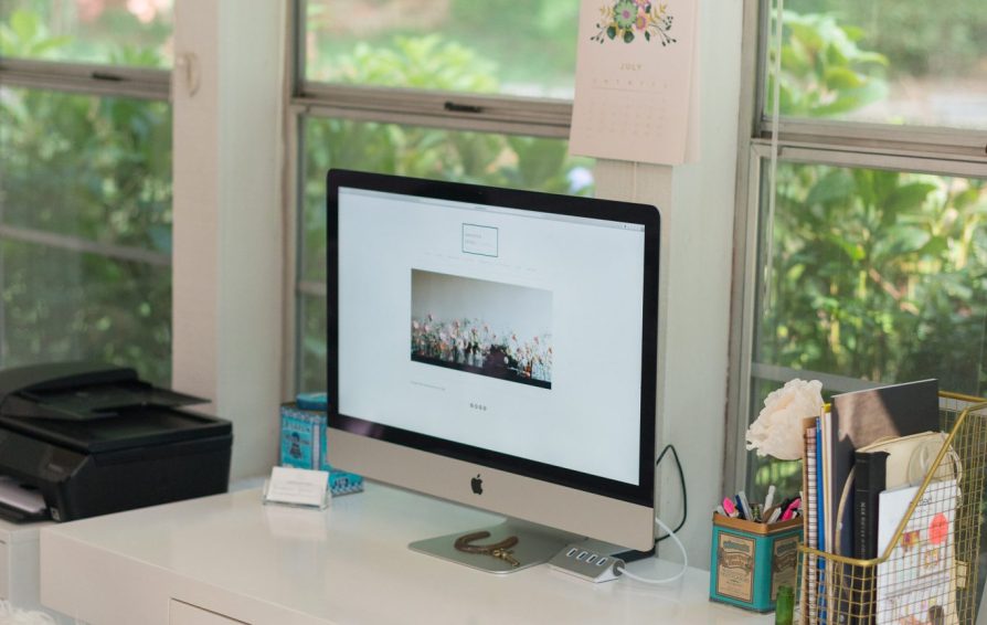 A white desk with an iMac in font of a wall of windows showing greenery outside.