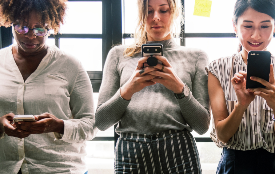 Three women stand side by side in front of windows, looking at their phones