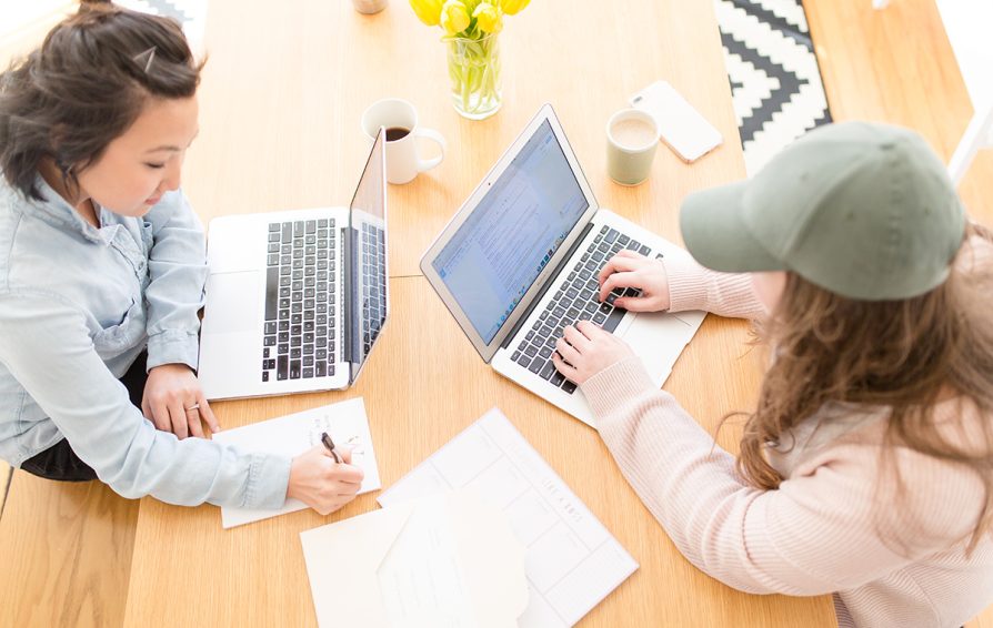 Aerial view of two women working on laptops at a wooden table.