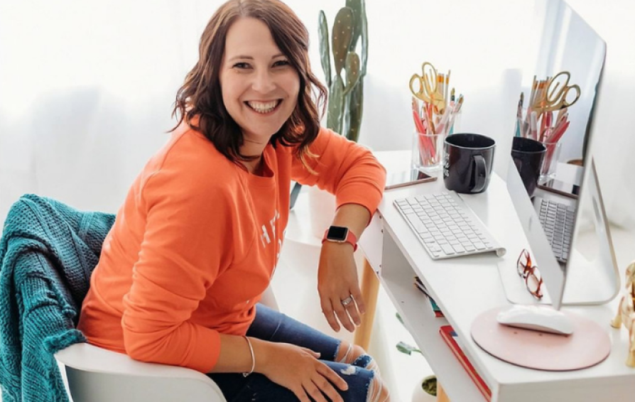Woman in an orange sweater at desk smiles over her shoulder.