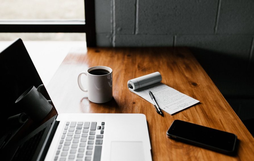 A laptop sits open on a wooden desk with a mug of coffee, notepad, and phone