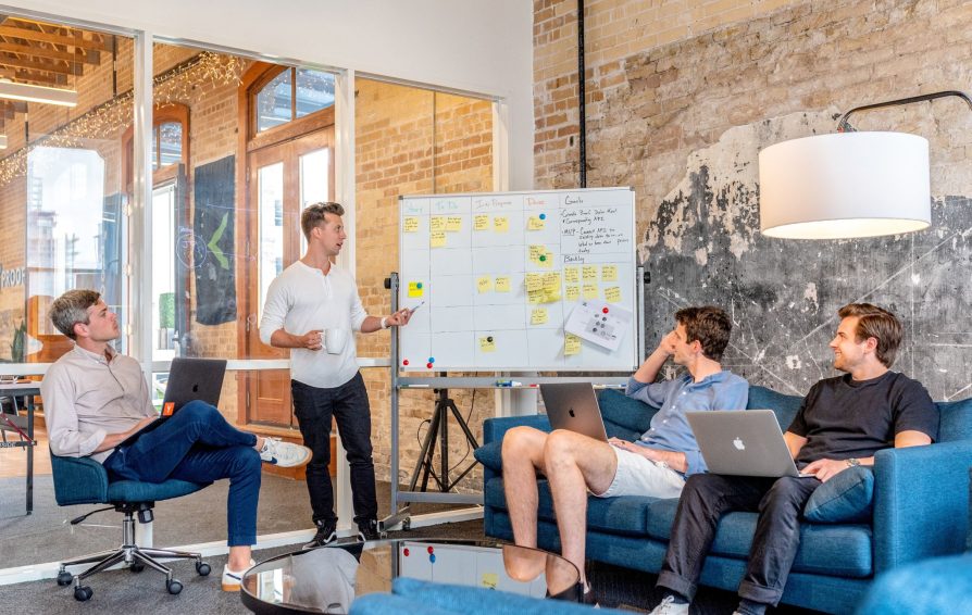 A man stands next to a whiteboard, giving a presentation to three men working on laptops.