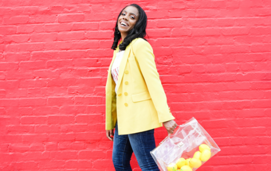 a smiling woman in a yellow blazer in front of a red brick wall carrying lemons