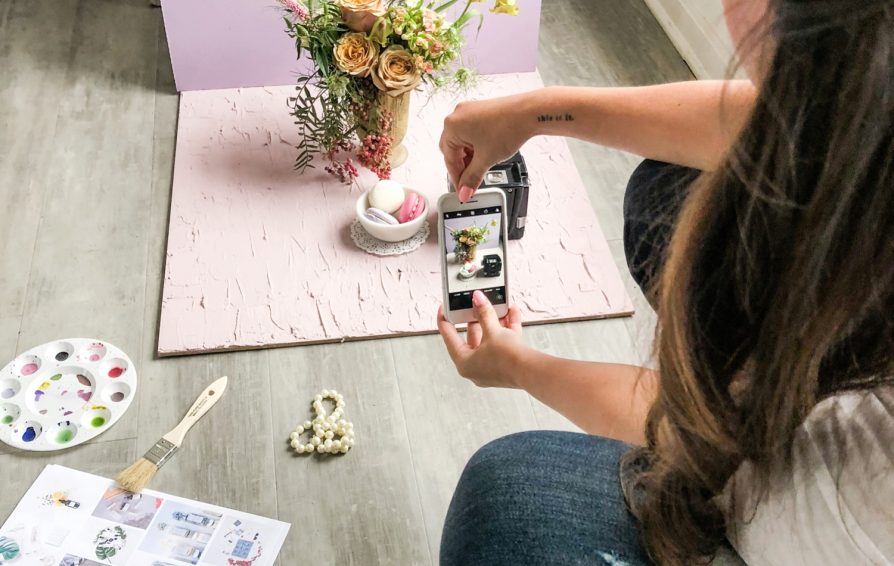 A woman takes a picture on her phone of a bowl of macarons, styled with a vase of flowers in a mini photo studio