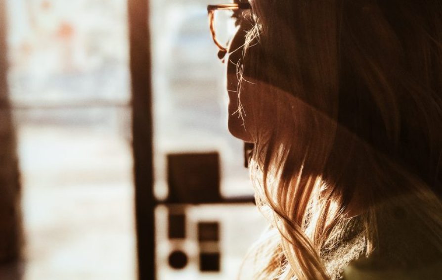 Woman with glasses looking out of a sunny window.