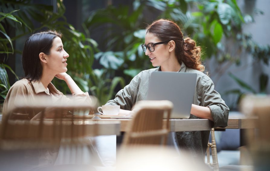 Content confident young ladies in casual shirts sitting at table in cafe and using laptop while working on web design Confident ladies working on web design
