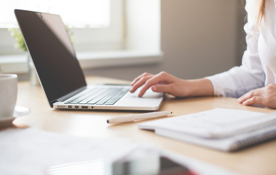 A person works at an open laptop on a desk with notebooks and coffee cup