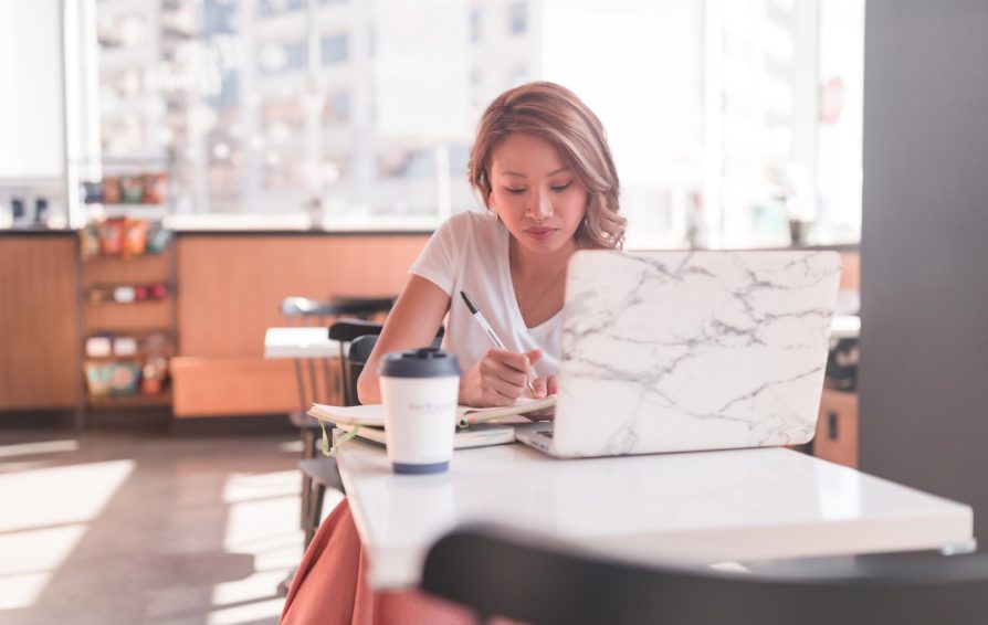 A woman sits at a table in a coffee shop, working on her laptop and writing in a notebook