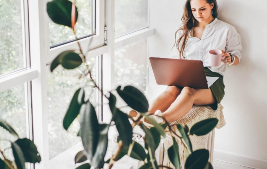 A woman sits in front of a large window, holding a mug with a laptop on her legs. There is a plant in the foreground.