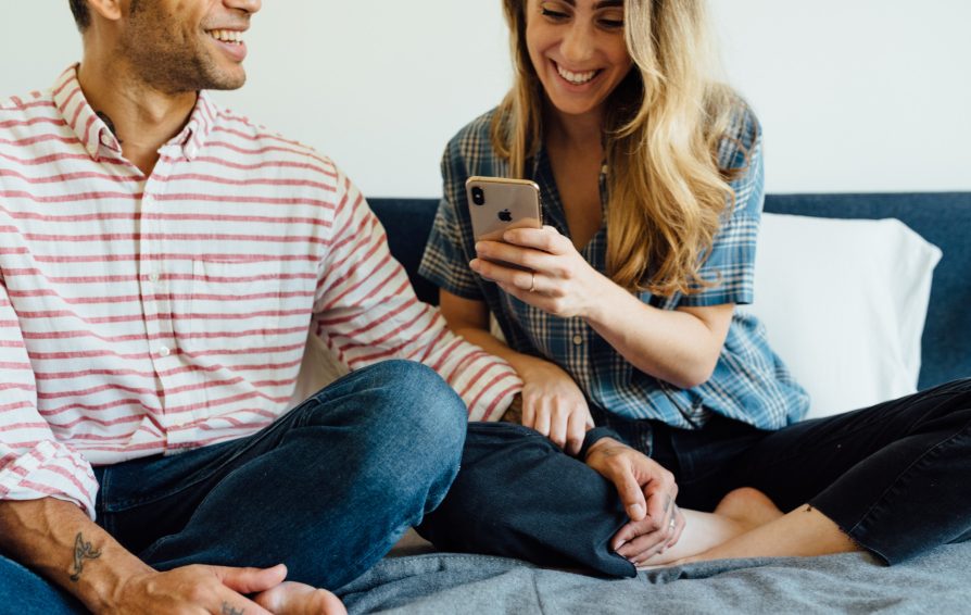 A man and woman sit cross-legged together on a bed, affectionately. The woman smiles at her phone in hand.
