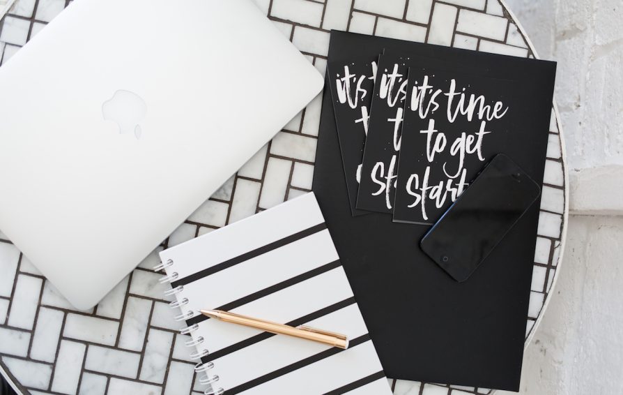 A white mosaic tile table with black grout is set with an iMac, a notebook, and folder with cards.