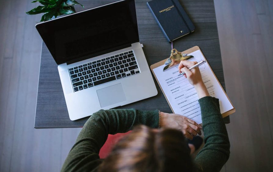 Aerial view of a person working at a table, writing on a clipboard. Beside them is an open laptop