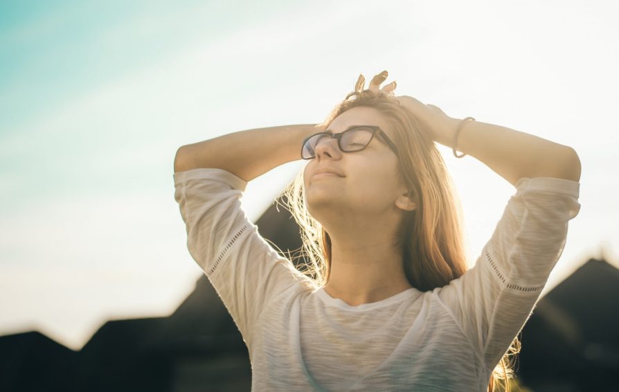 A woman wearing glasses basks in the sun with hands on her head
