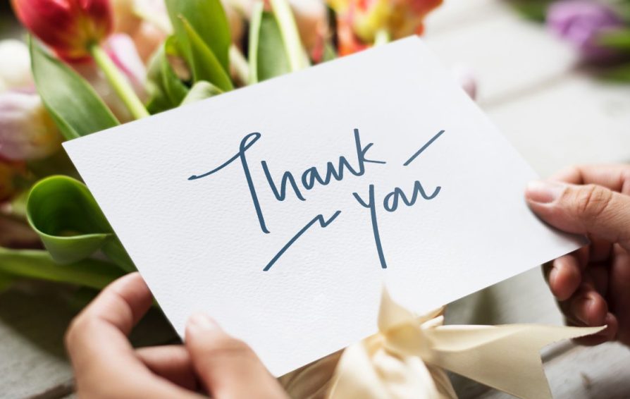 A pair of hands hold a thank you card in front of a bouquet of flowers