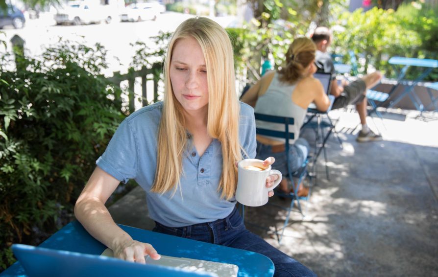 A woman sits at a park, holding a mug of coffee and working on her laptop at a table