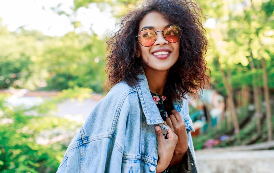 Close up of a woman smiling outdoors. She holds a denim jacket draped over her shoulders.