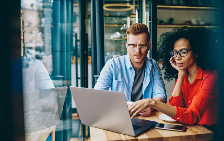 A man and woman work on a laptop next to a large window