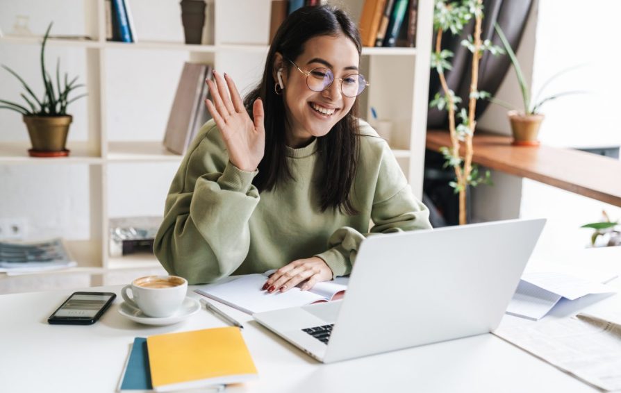 a woman with glasses sitting at a desk waving to her laptop