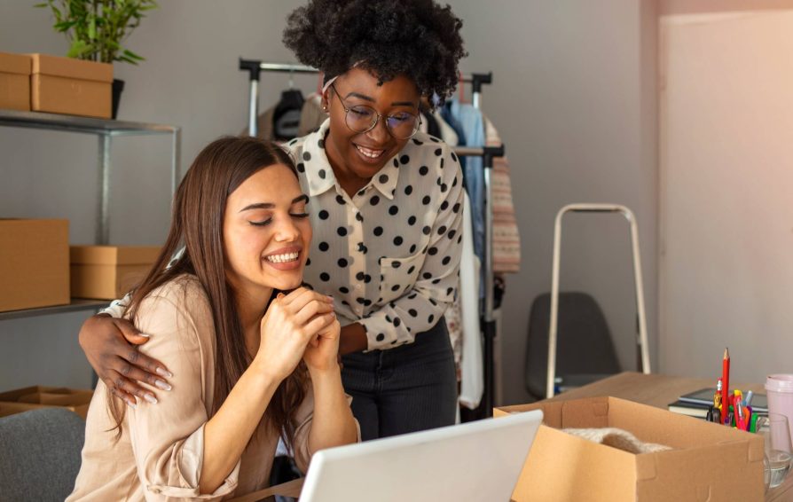 A woman sits at a desk with another woman standing beside her with arms around her shoulders. They both look happily at the computer on the desk.