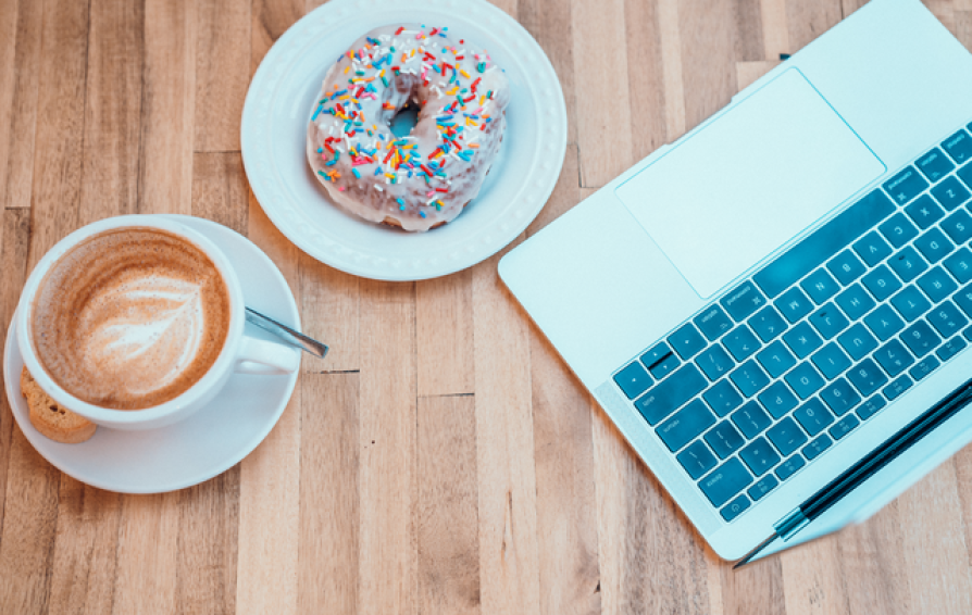 Aerial view of a latte, a donut, and a laptop on a wooden surface.