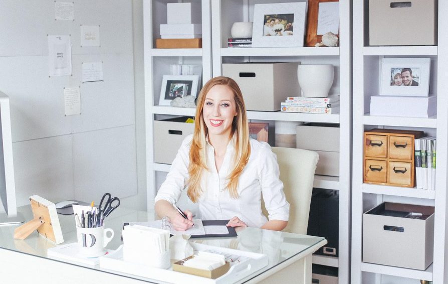 A woman sits smiling at a white desk. The office is light with styled white shelves in the background