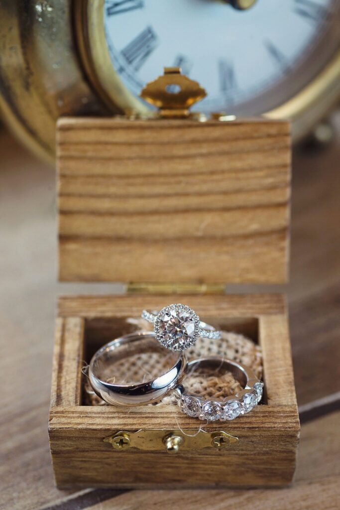 Silver and diamond wedding rings sit in a wooden ring box with a clock in the background