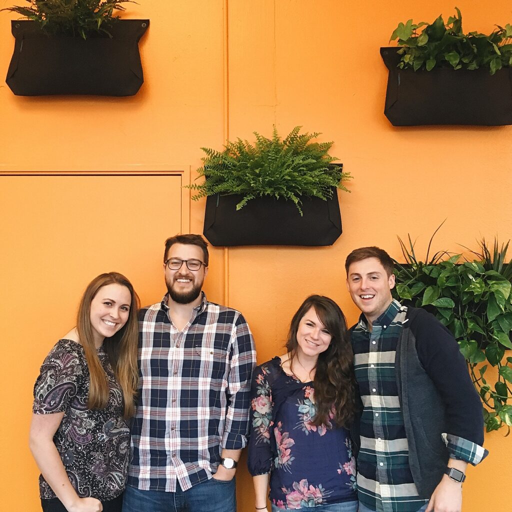 A group of people stand in front of an orange wall with plants, smiling at the camera.