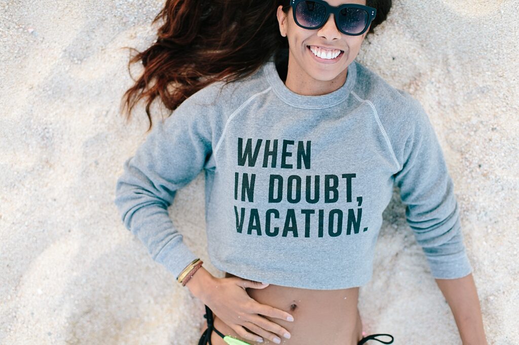 Aerial view of a woman lying in the sand, smiling. She wears sunglasses and a sweatshirt that says 