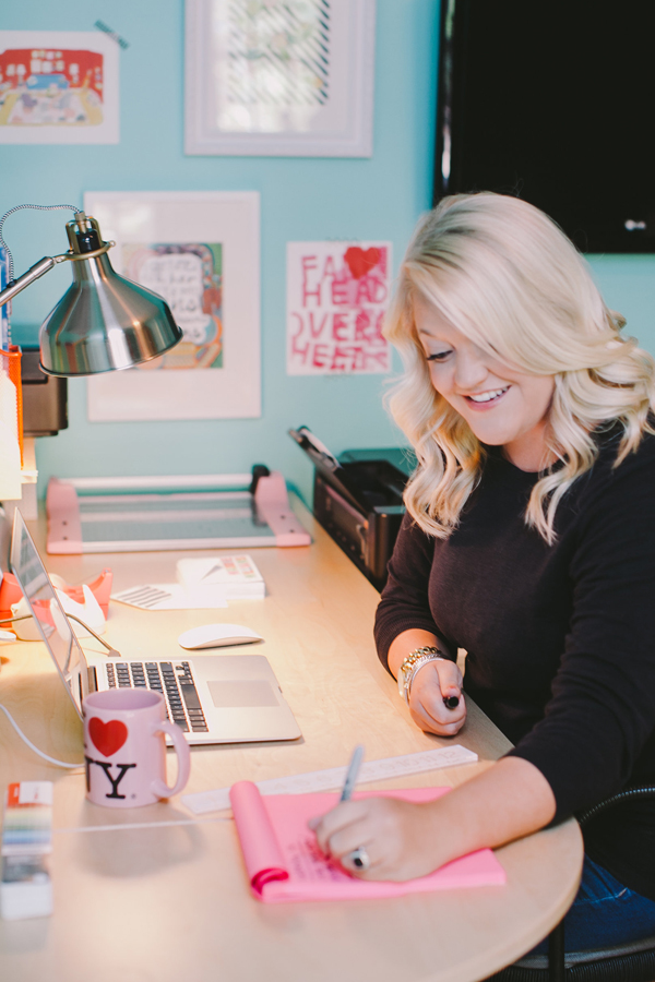 A blonde woman sits, smiling at a desk as she writes a list on pink paper