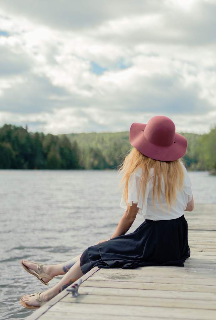 A woman wearing a pink hat sits on a dock, looking out towards the water