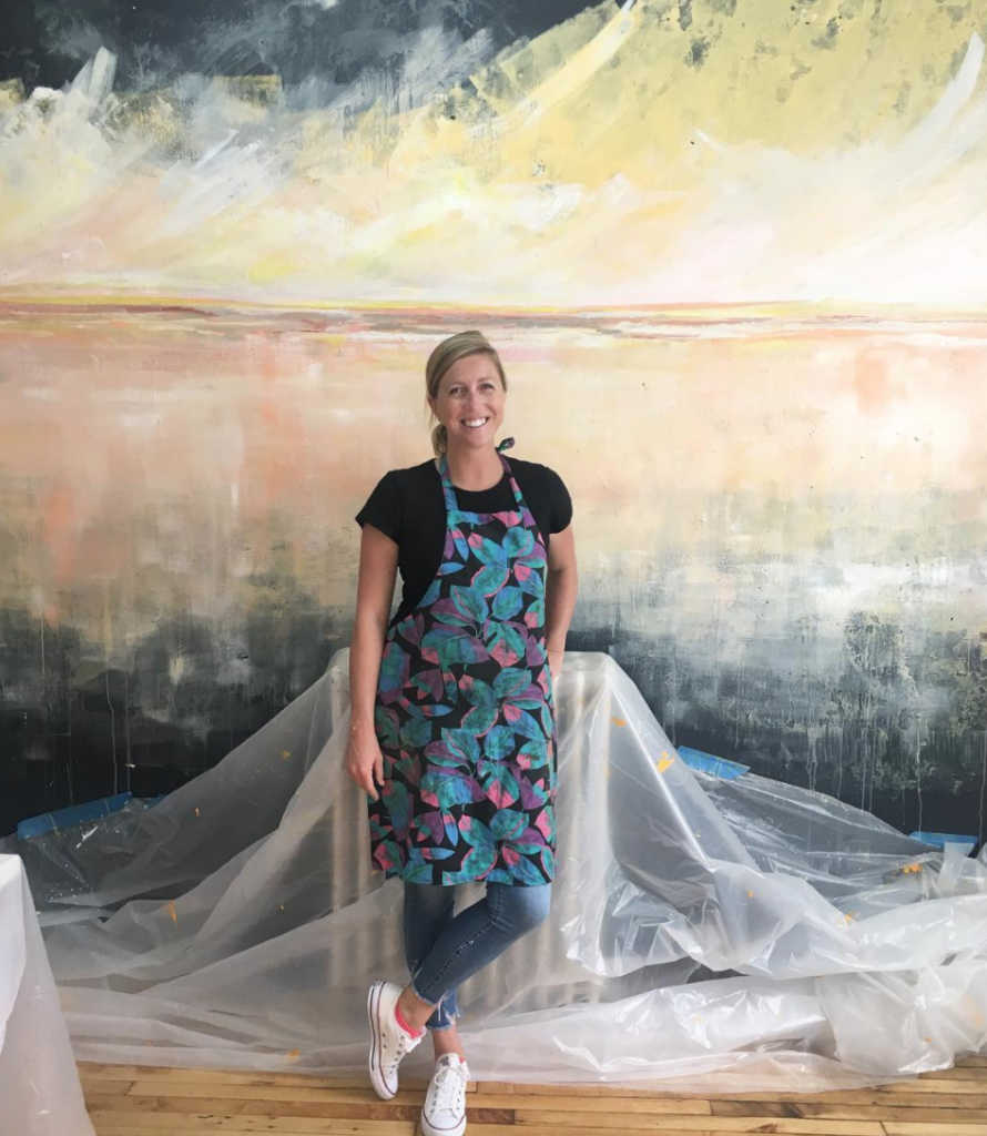 A woman in an apron stands in front of a large, abstract painting.