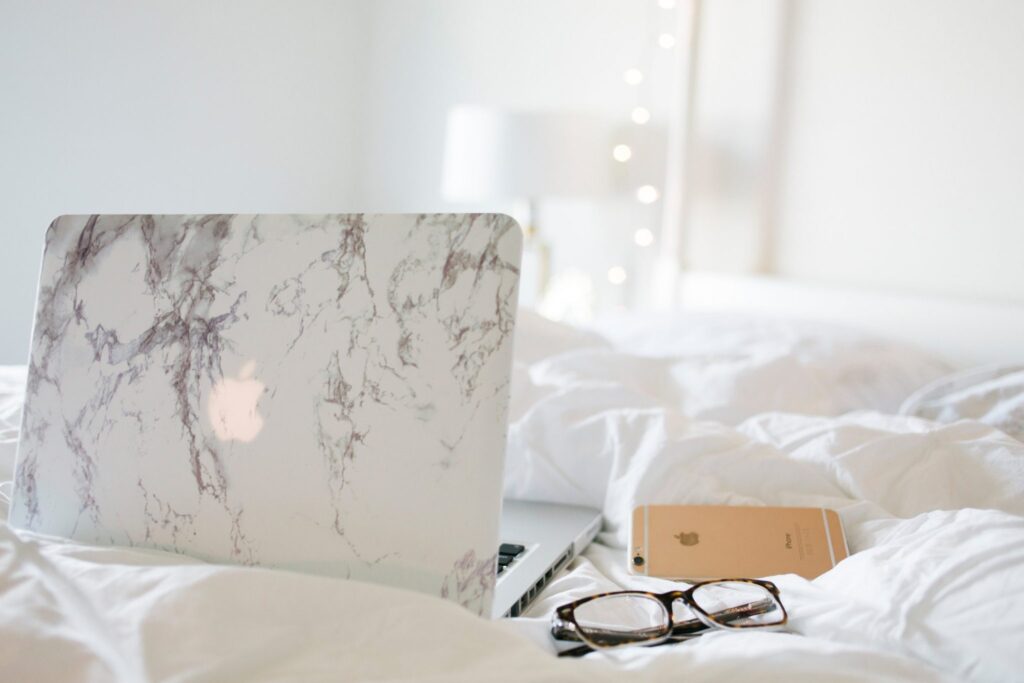 A Macbook with a marble cover, pair of glasses, and iPhone sit on a fluffy, white bed.