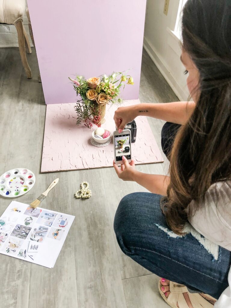 A woman takes a picture on her phone of a bowl of macarons, styled with a vase of flowers in a mini photo studio