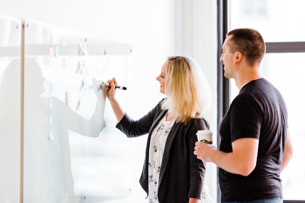 A woman draws on a whiteboard while a man holding a coffee looks on
