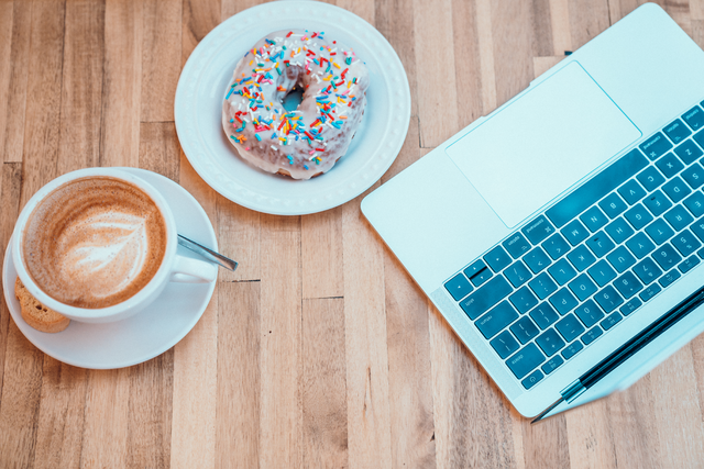 Aerial view of a latte, a donut, and a laptop on a wooden surface.