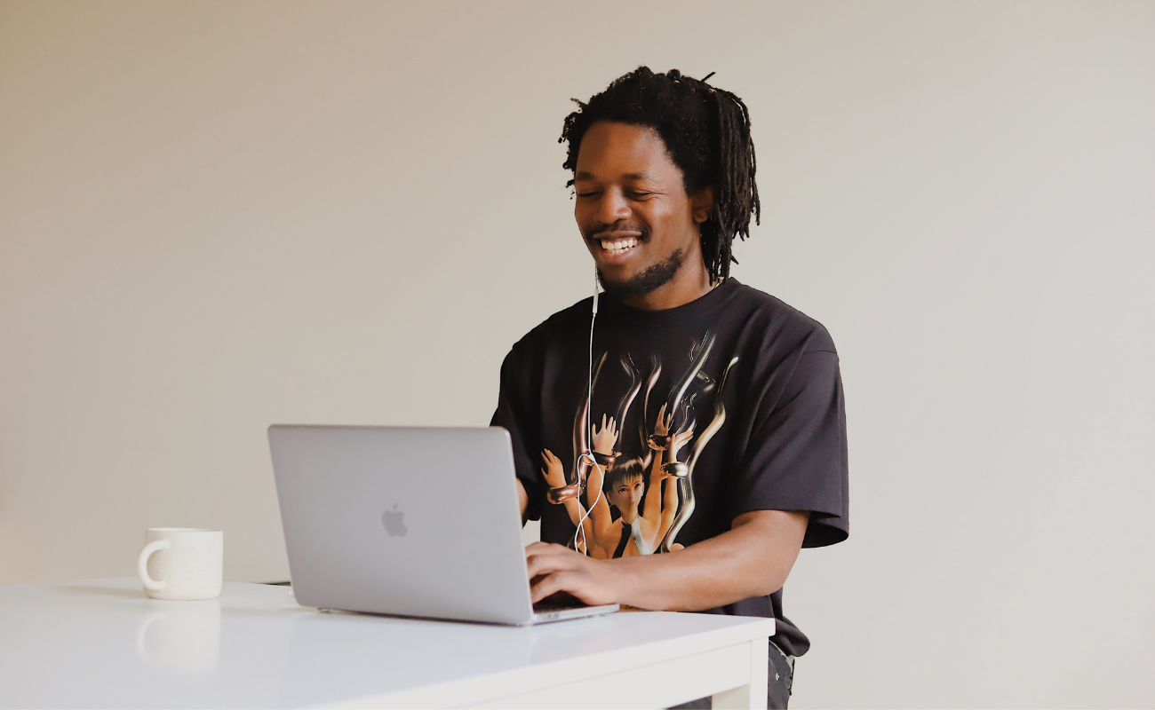 A black man sitting at a white table with a laptop open and coffee mug next to it looks down and smiles