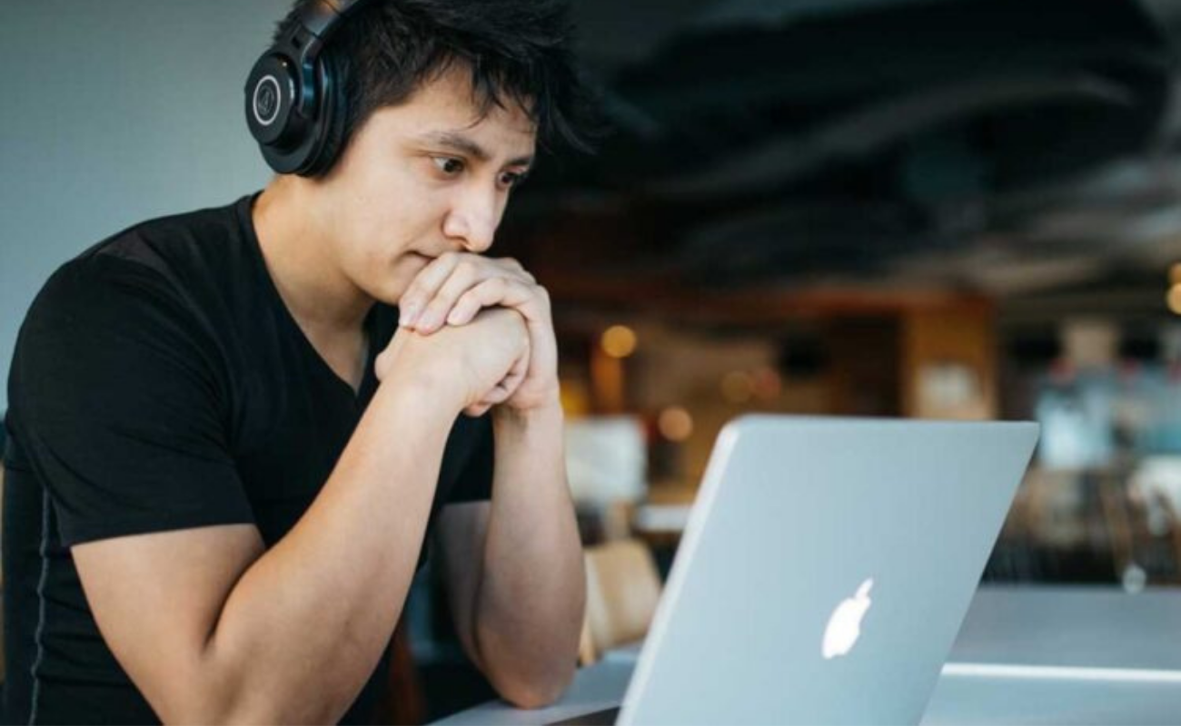 A man sits at a desk with headphones on looking down at a laptop with his hands on his chin
