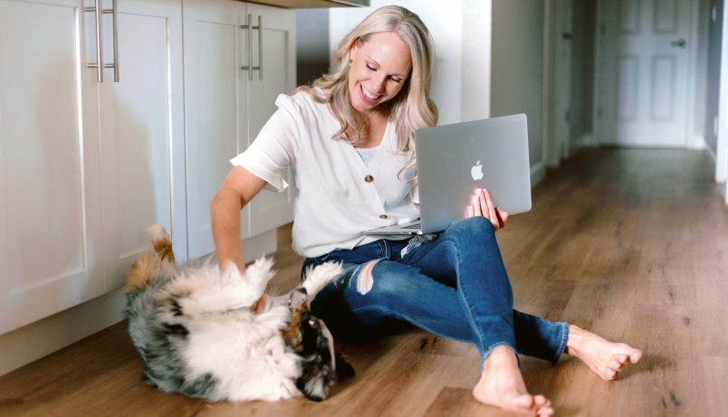 a woman sitting on the floor holding her laptop and petting a dog