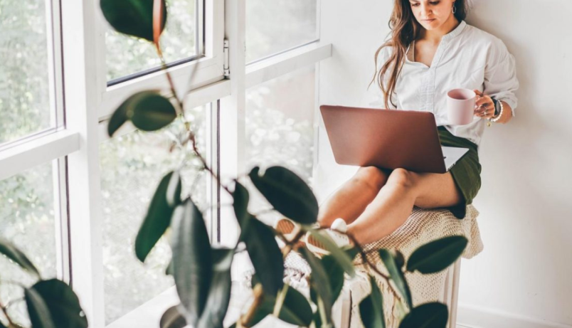 A woman sits in front of a large window, holding a mug with a laptop on her legs. There is a plant in the foreground.
